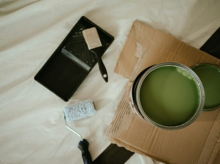 An open can of green house paint beside a paint tray and brush on a protective sheet.